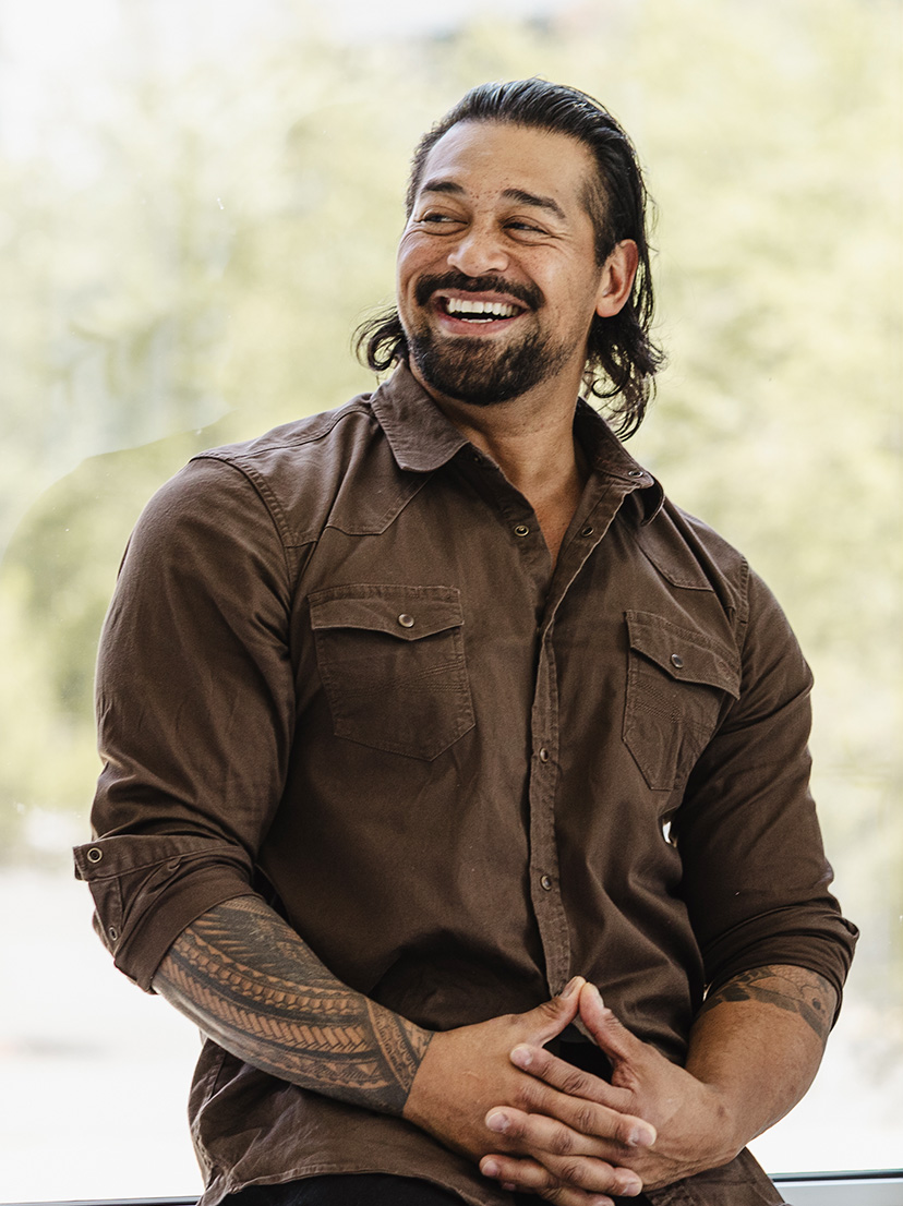A man in his forties in a brown shirt, laughing, hands clasped, outdoors
