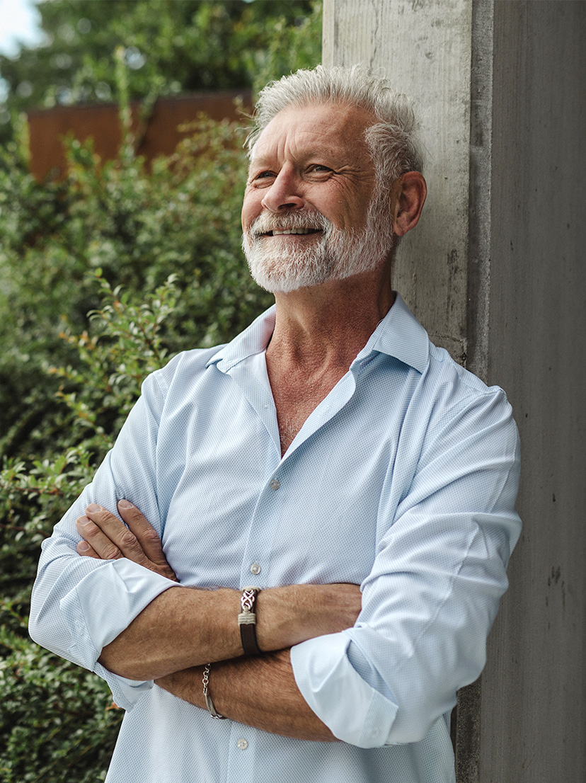 An older man with white hair and a beard, arms folded, smiling outdoors