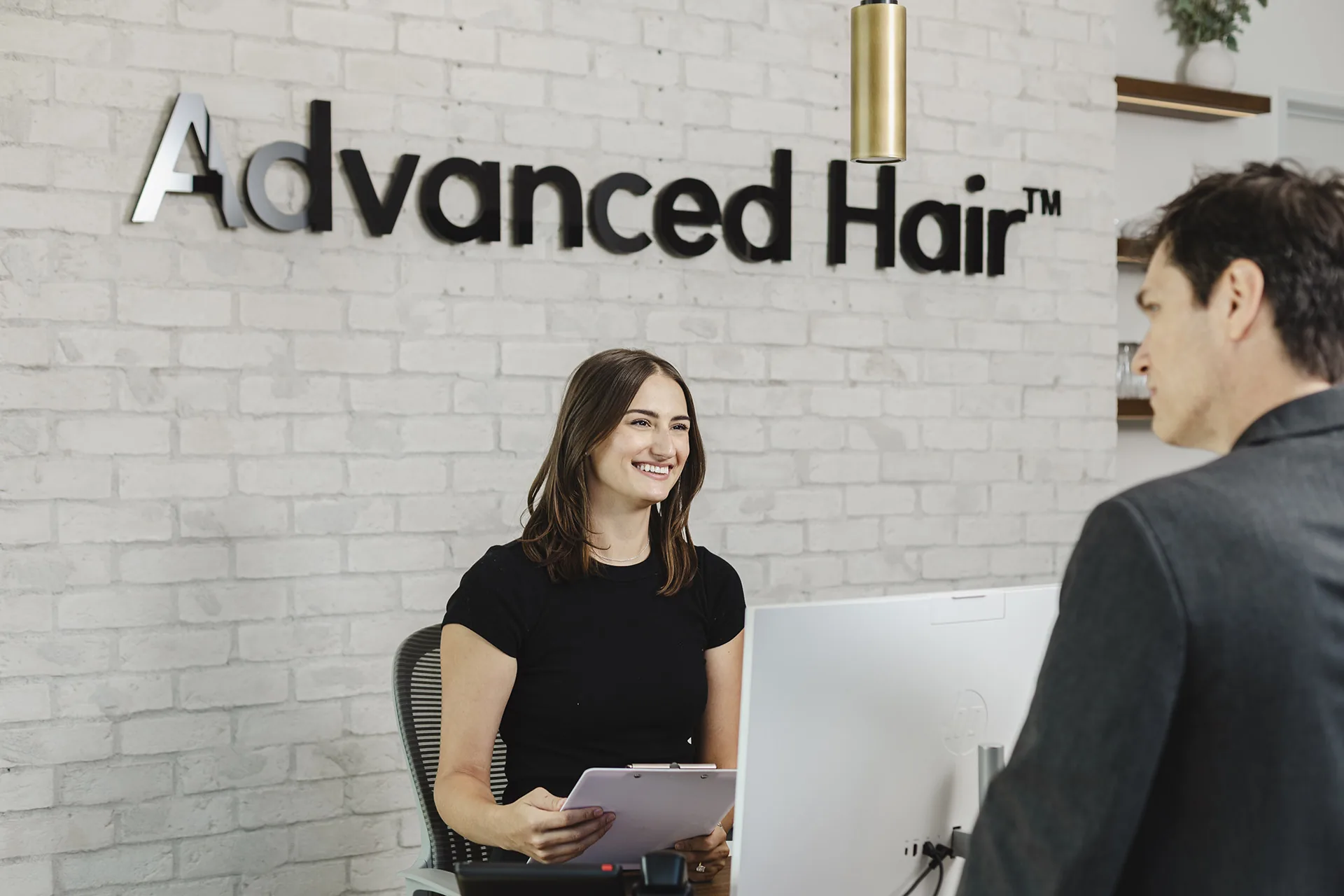 A receptionist smiling at a visitor across the front desk at Advanced Hair Restoration, the clinic logo mounted on the white brick wall behind her