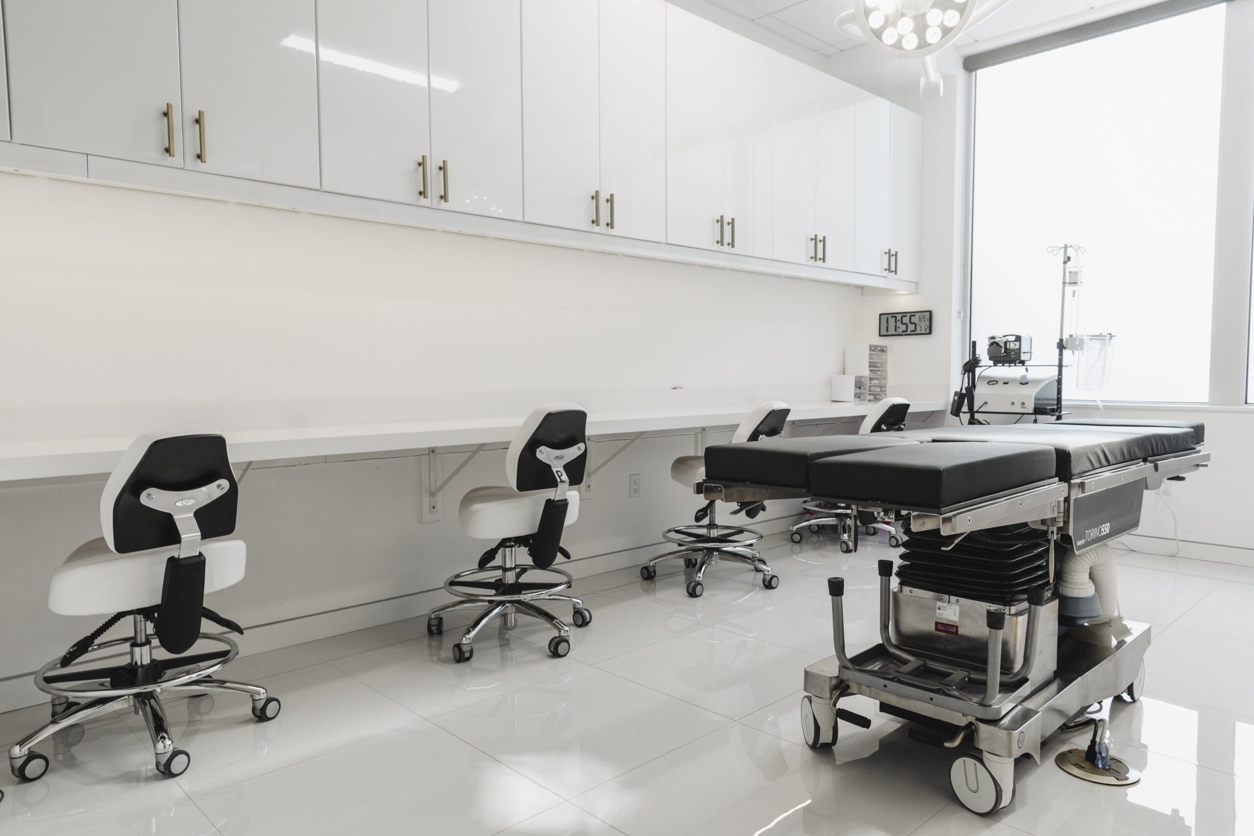 A white surgical suite with a procedure table, surgeon stools and instrument monitors beside a bright window