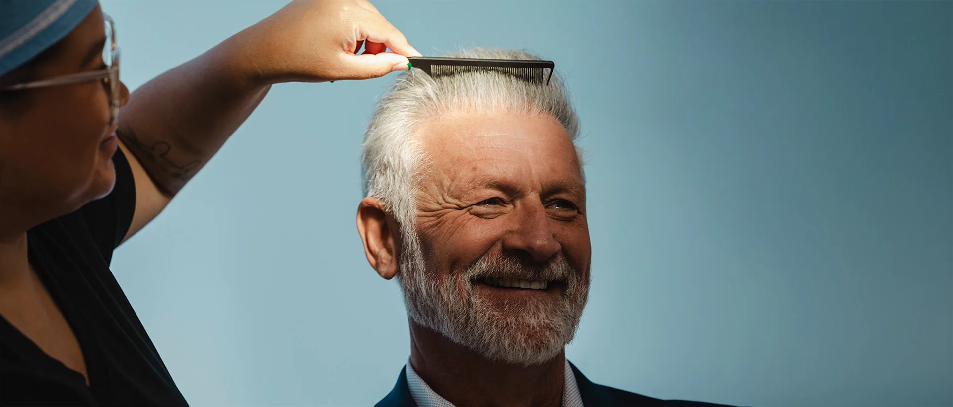 A clinician in scrubs combing the hair of a smiling older man against a blue backdrop