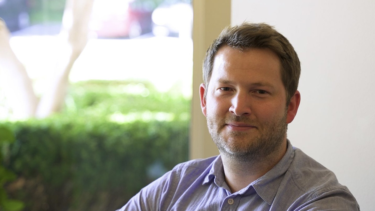 Brendan Hay smiling gently indoors, beside a window with greenery outside